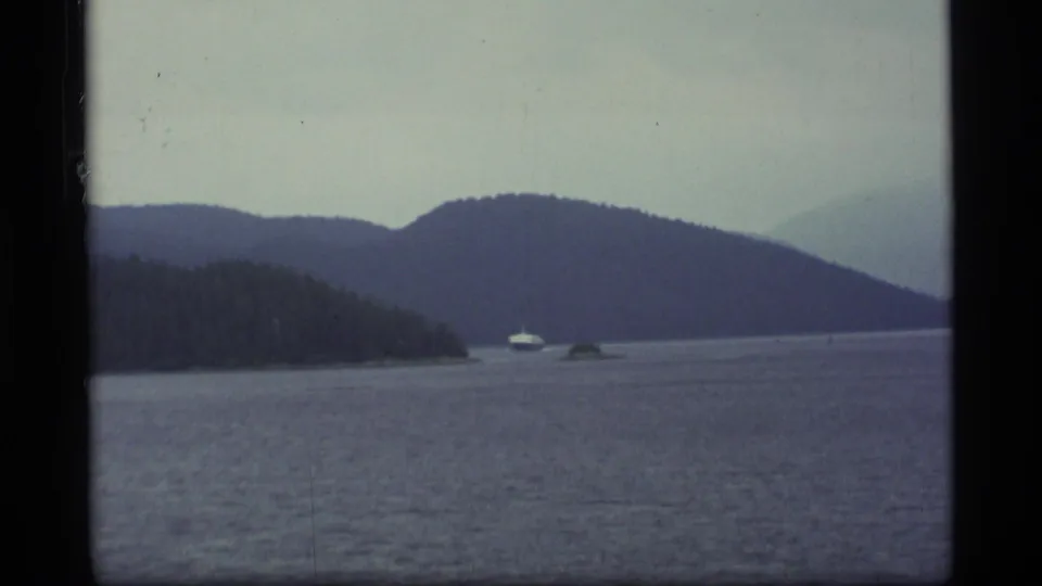 1977 Alaska 8mm frame shows a small white boat near forested shoreline with broad water and hazy mountains beyond.