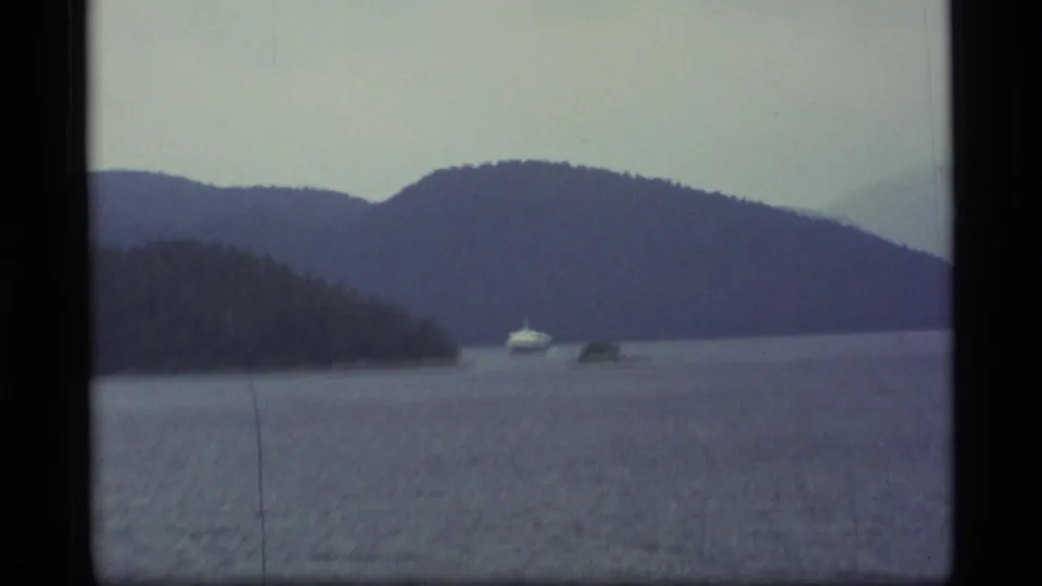 1977 Alaska 8mm frame showing a distant boat crossing calm water between evergreen islands and layered mountain ridges.