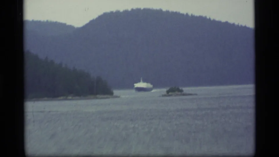 1977 Alaska home-movie frame with a large boat near forested islands, backed by blue mountain slopes and overcast sky.