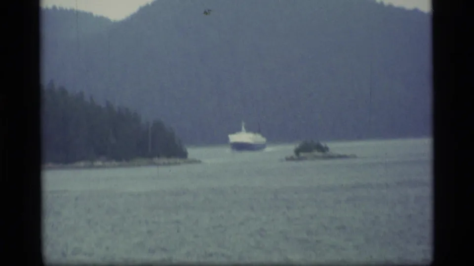 1977 Alaska 8mm view of a boat crossing a broad lake near a tree-covered island, with mountain terrain in the background.