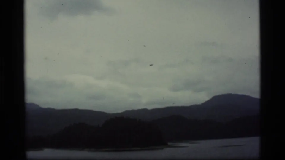 1977 Alaska 8mm frame showing a dark lake shoreline under low clouds, with mountain ridges and a small distant speck that may be aircraft.