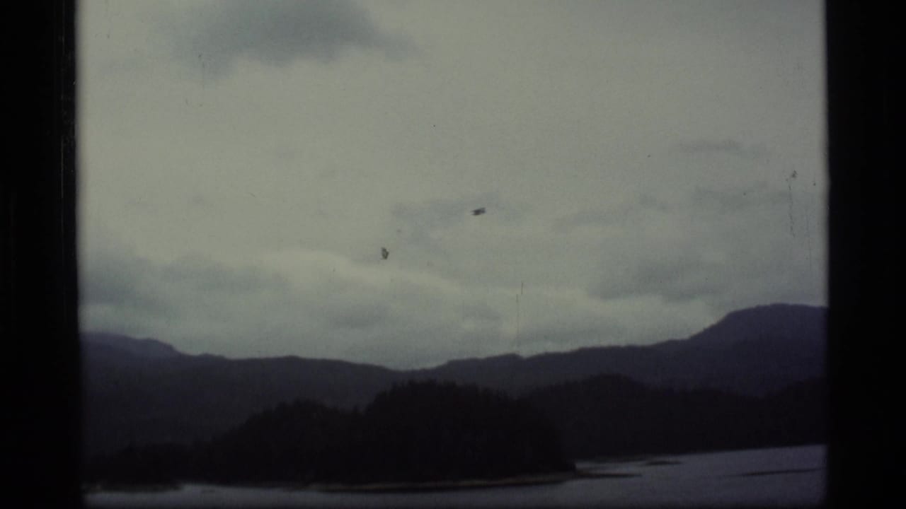 1977 Alaska 8mm frame showing distant mountains, a lake, and heavy clouds before the seaplane becomes prominent.