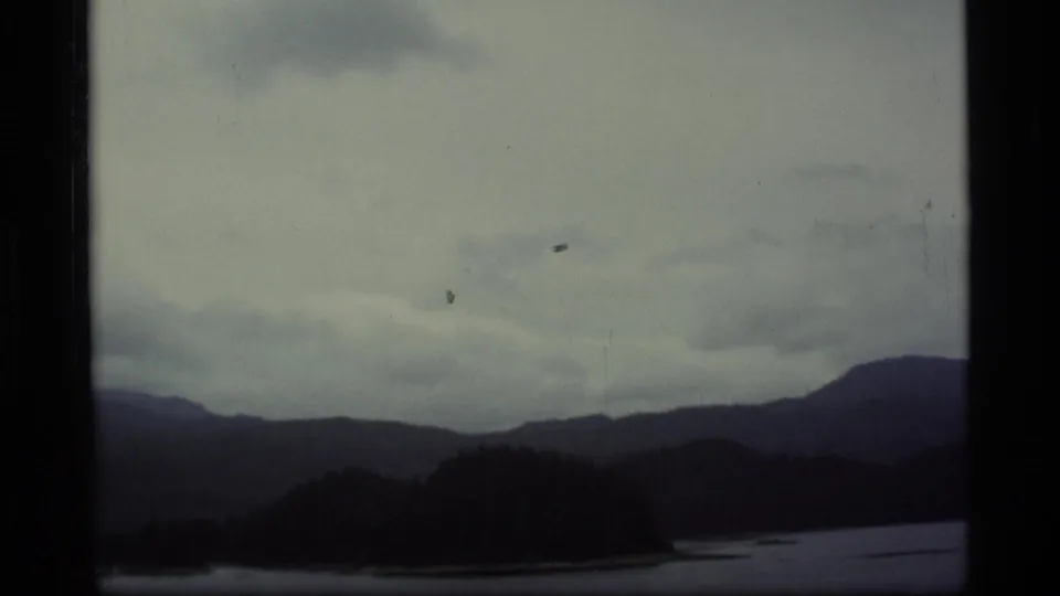 1977 Alaska 8mm frame showing distant mountains, a lake, and heavy clouds before the seaplane becomes prominent.
