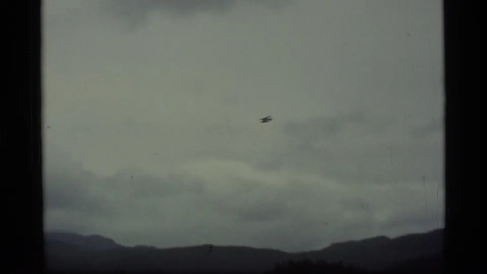 Small seaplane appears against an overcast Alaska sky, with low mountains along the bottom of the frame.