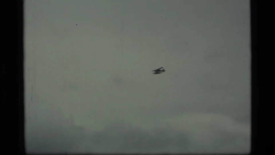 1977 Alaska home-movie frame of a small seaplane seen in profile against muted cloud cover.