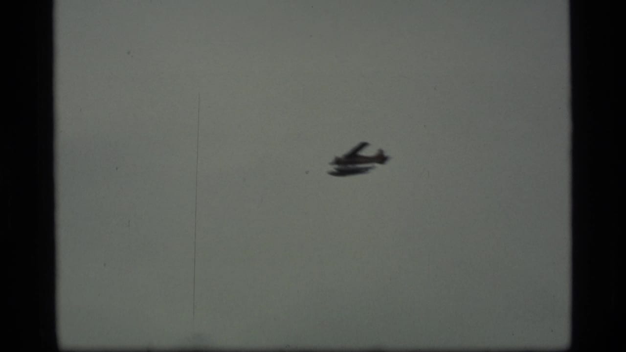 Small propeller plane appears against a gray sky in 1977 Alaska home-movie footage, with visible film borders and scratches.