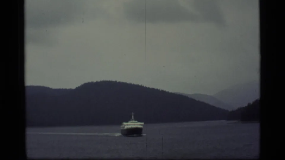 A ferry moves through an Alaskan waterway, with dark wooded hills and layered mountains visible beyond the water.