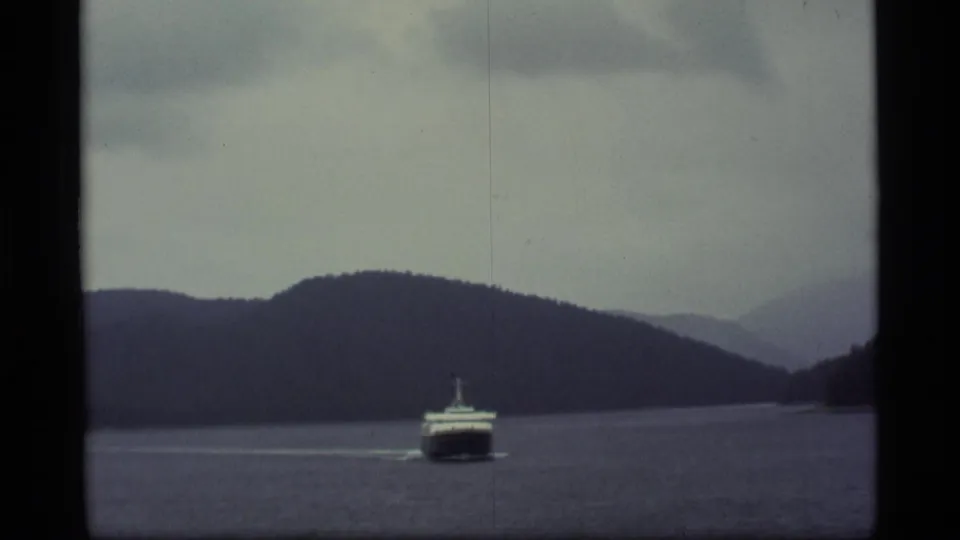 The ferry continues across the waterway in 1977 Alaska footage, framed by dark hills, pale clouds, and worn 8mm film edges.