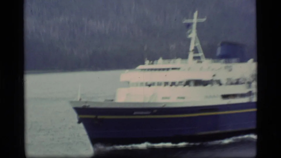 1977 Alaska frame of a blue-and-white ferry moving across calm water below a wooded hillside.