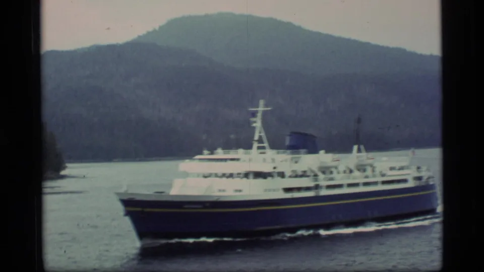1977 Alaska home-movie frame with a passenger ferry, calm water, wooded shore, and rounded mountain backdrop.
