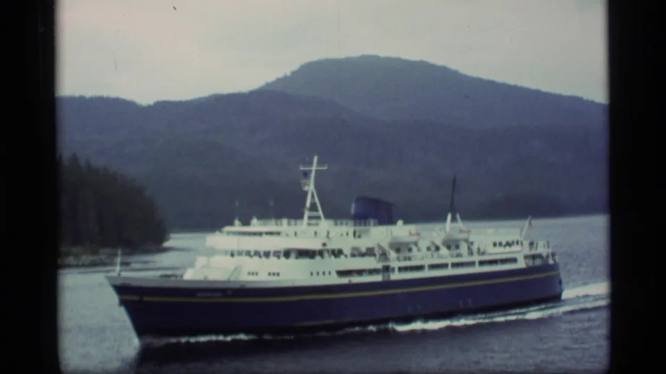 1977 Alaska frame showing a ferry in three-quarter view with passengers faintly visible and hills behind the waterway.
