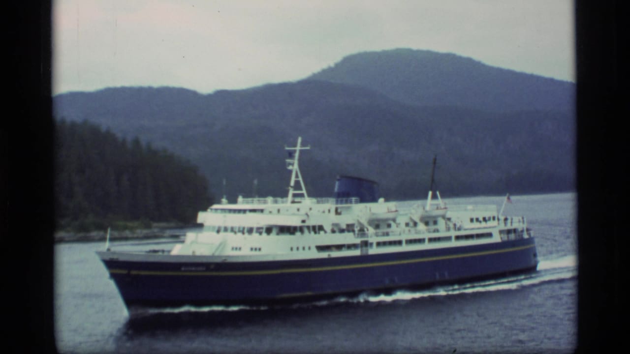 Blue and white ferry crossing calm water with forested shore and mountains behind it.