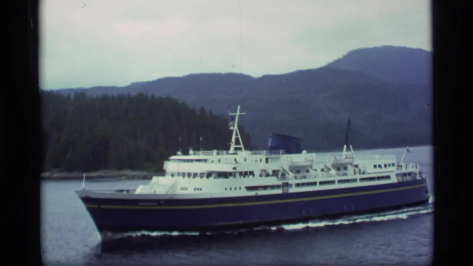1977 Alaska 8mm frame of a blue and white ferry traveling across lake water under an overcast mountain horizon.