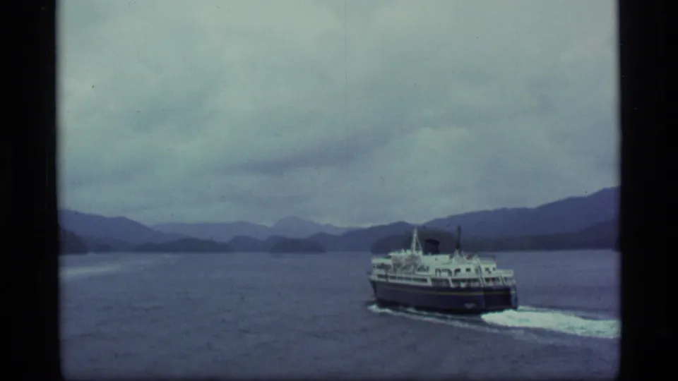 1977 Alaska 8mm frame showing the ferry receding across open water toward layered hills and distant mountains.