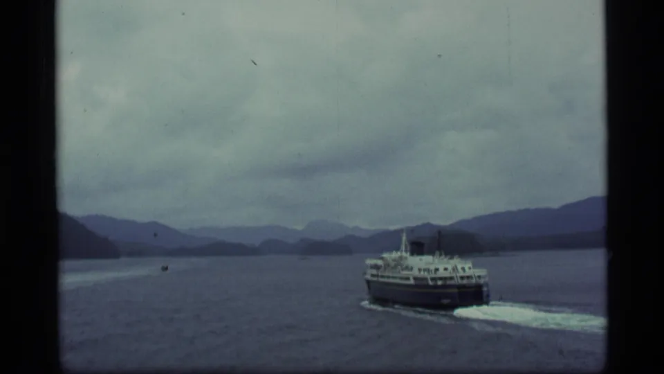 1977 Alaska 8mm frame of the ferry farther from camera, crossing broad water with mountains under a muted sky.