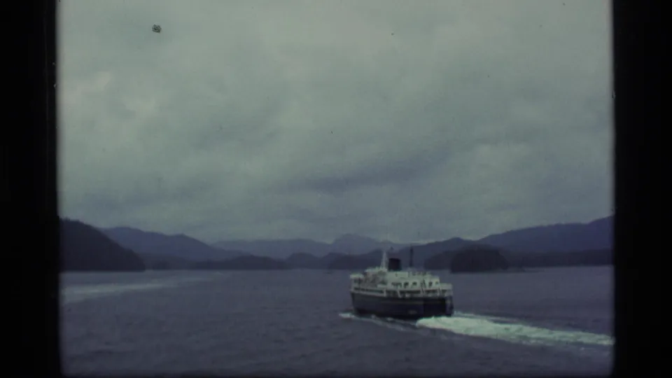 Vintage 8mm view of a ferry moving through an Alaskan waterway under cloudy skies and dark mountain ridges.