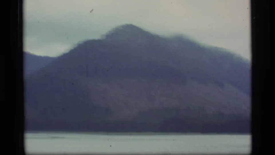 1977 Alaska home-movie frame with a soft-focus mountain slope rising above a pale waterline.