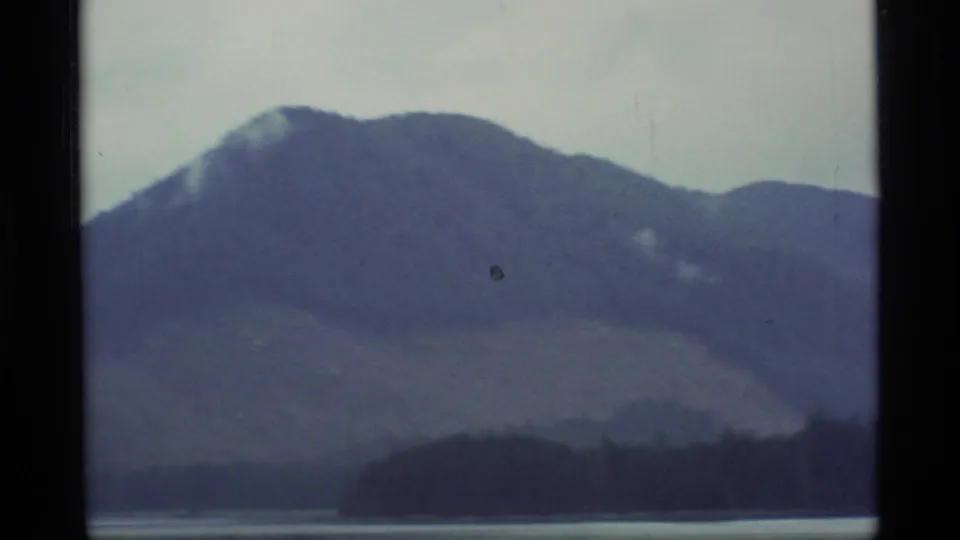 1977 Alaska home-movie frame with a broad misty mountain rising behind a low wooded shoreline and water.