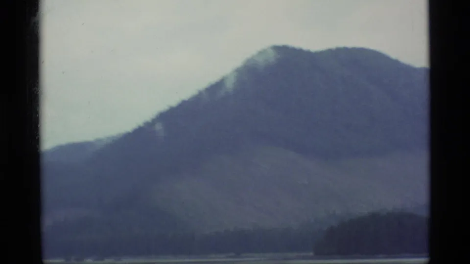 1977 Alaska 8mm frame with a soft, close view of a misty mountain slope and dark shoreline near water.