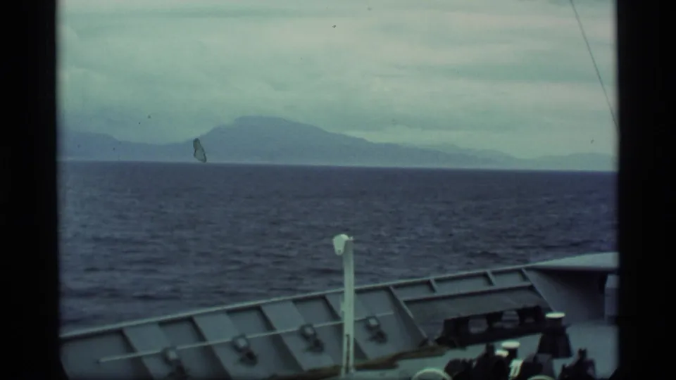 1977 Alaska home-movie frame from a vessel deck, looking over railing toward open water and distant hazy mountains.