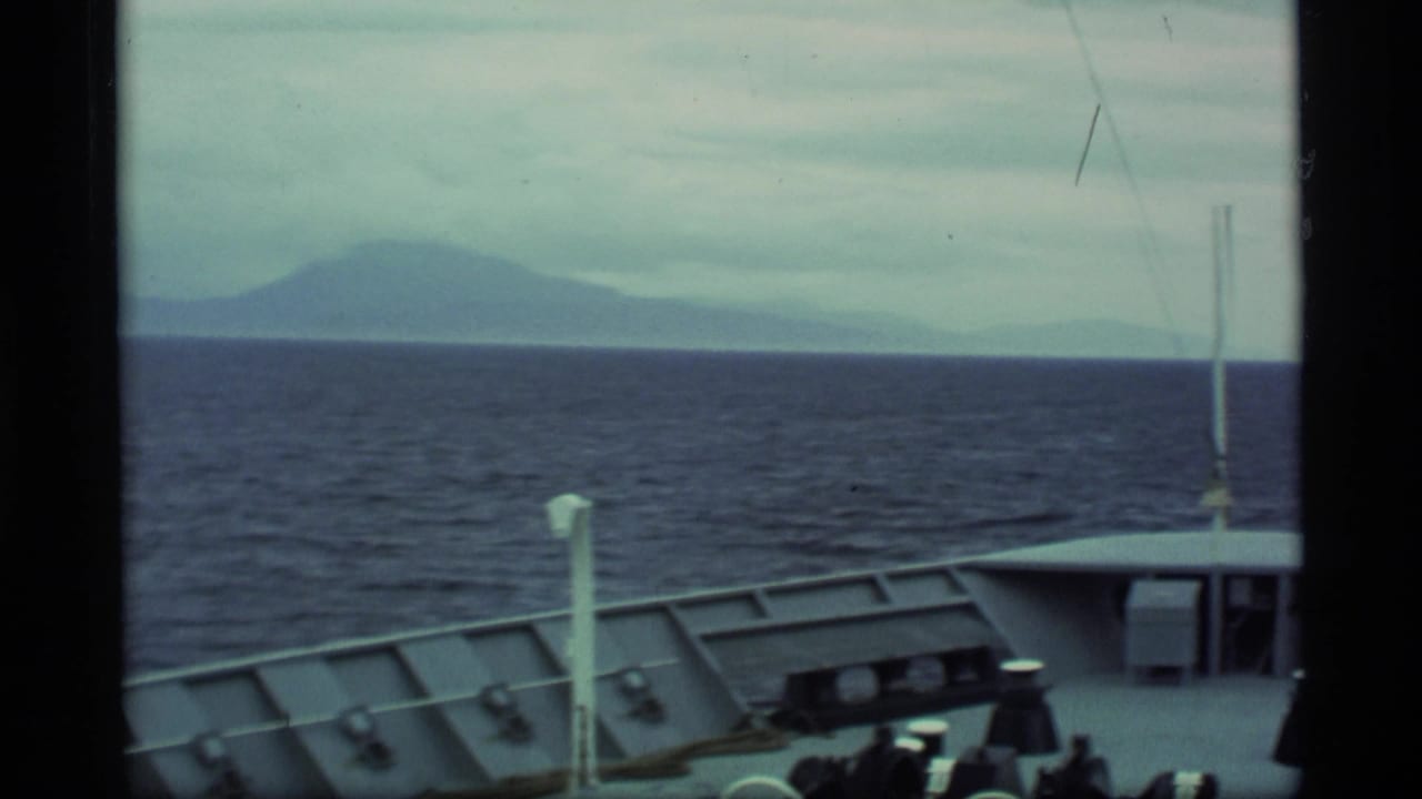 Ship bow and railings face open water with distant mountains under a cloudy sky.