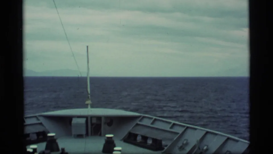 1977 Alaska 8mm frame with the ship bow low in view, a small mast, dark sea, and distant coastal mountains.