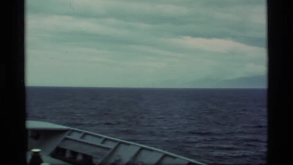 1977 Alaska cruise footage showing mostly open water, cloudy sky, and a partial bow with faint mountains on the horizon.