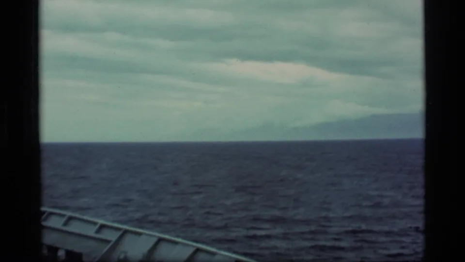 1977 Alaska home-movie frame showing open water, overcast sky, distant land, and a sliver of boat structure in the foreground.