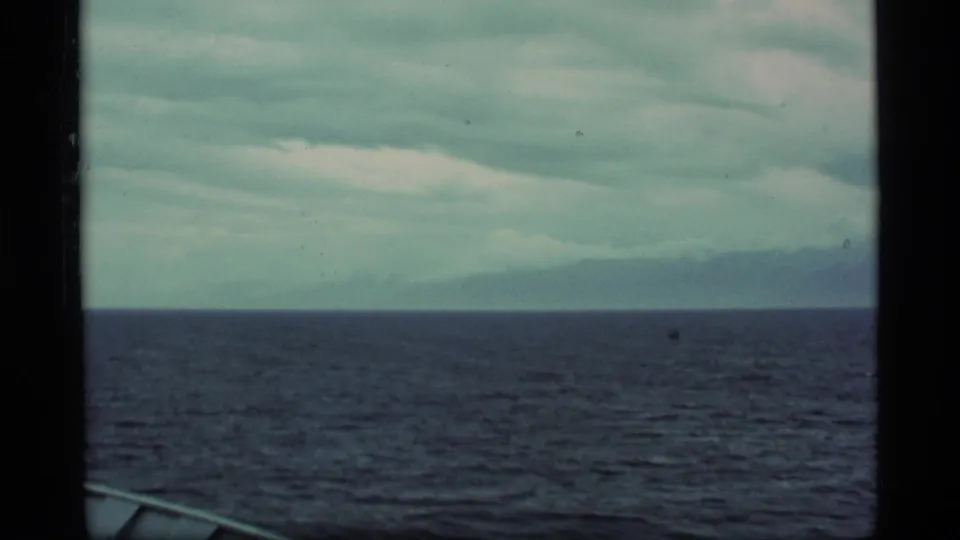 A 1977 Alaska travel frame looking across dark water toward a low, distant coastline beneath overcast clouds.
