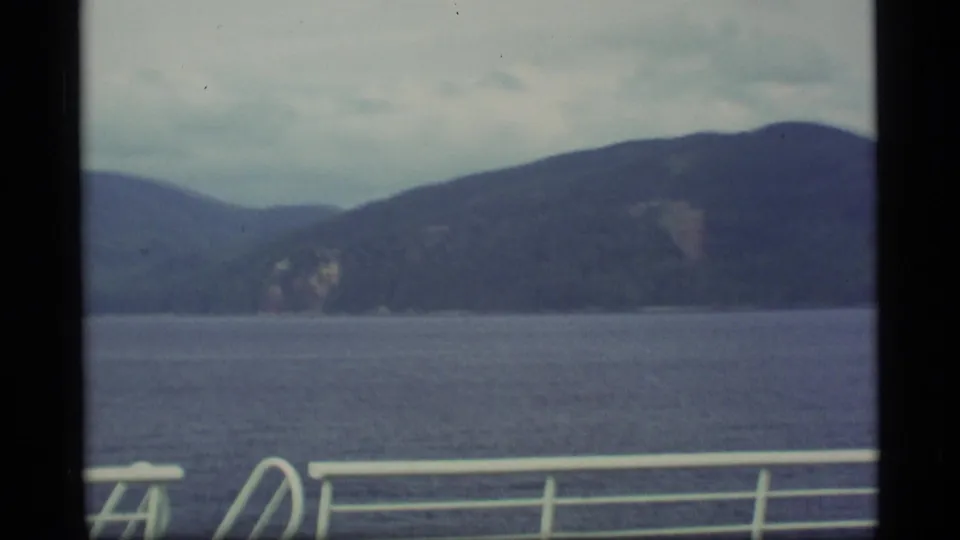 Alaska coastline and mountains seen across calm water from a boat deck, with white railing in the foreground.