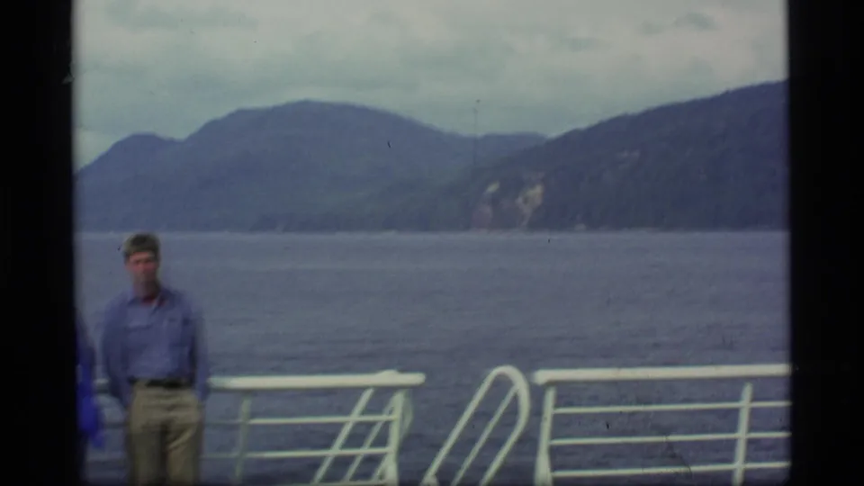 A passenger stands near the white railing on a boat deck, with Alaskan water and forested mountains in the background.