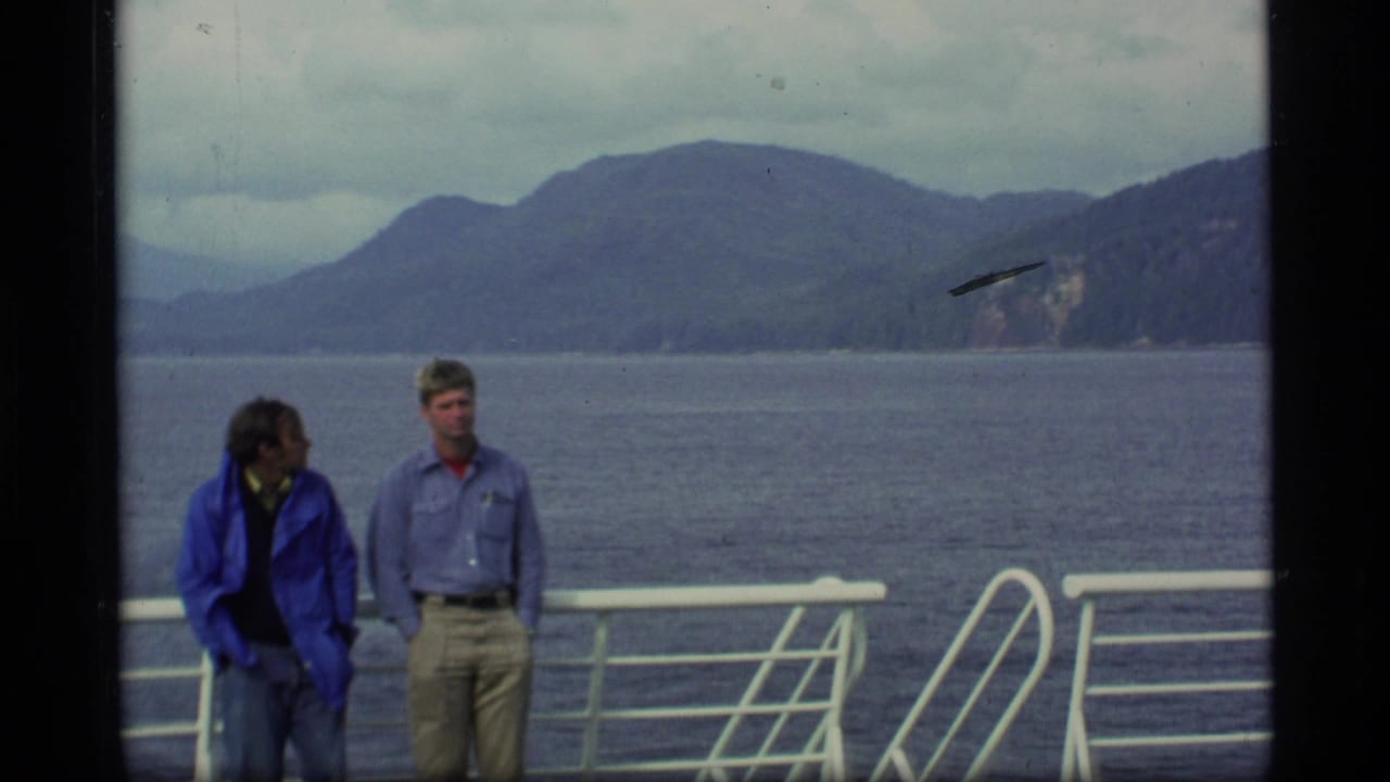 Two adult men on a ship deck in Alaska, with white railings, open water, and wooded mountains in the distance.