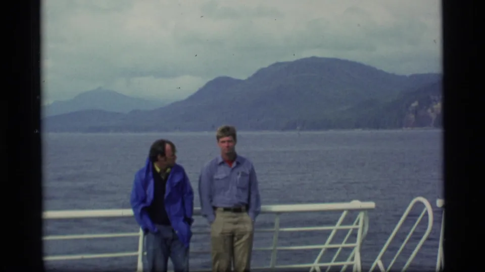 Two adult men on an Alaska ship deck, framed against calm water and dark mountain slopes under cloudy sky.