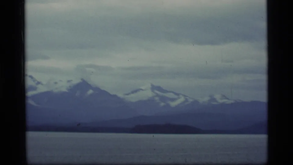 Alaska mountain landscape with snow-capped peaks, dark shoreline, water, and an overcast sky in soft 8mm color.
