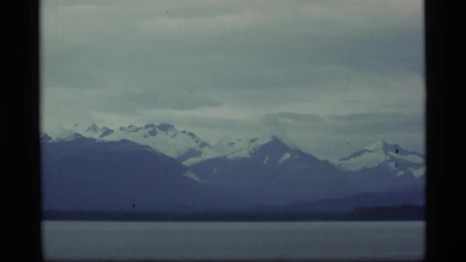 Wide Alaska view of snow-covered mountain peaks across water, with layered ridges and cloudy sky on 8mm film.
