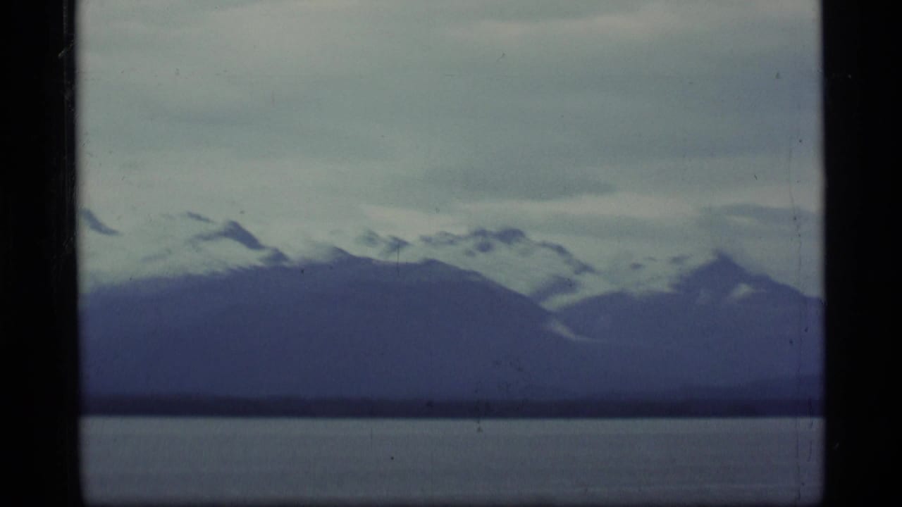 1977 Alaska 8mm frame showing snow-capped mountain ridges beyond a calm lake beneath a gray, clouded sky.