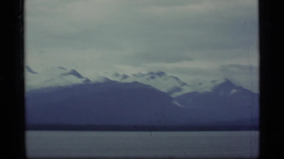 1977 Alaska 8mm frame showing snow-capped mountain ridges beyond a calm lake beneath a gray, clouded sky.