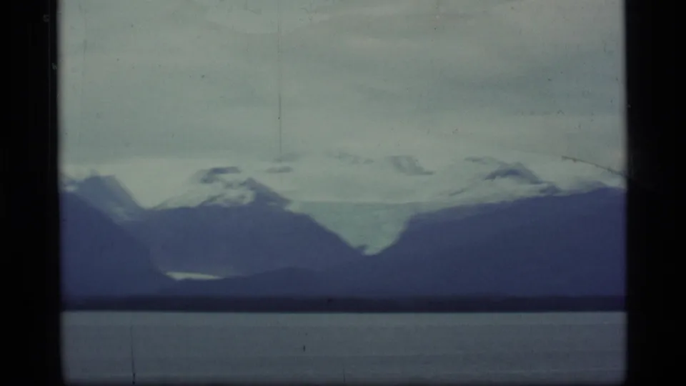 1977 Alaska 8mm frame with distant snowy peaks, dark foothills, and a calm lake in the foreground.
