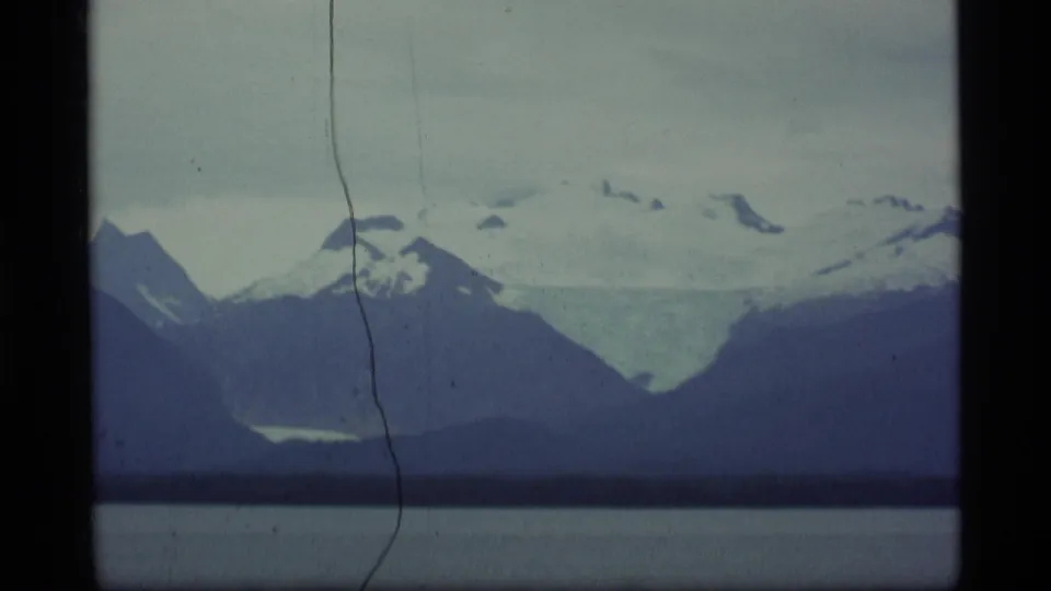 1977 Alaska 8mm frame showing snow-patched mountains over a lake, with scratches and gate edges visible in the scan.