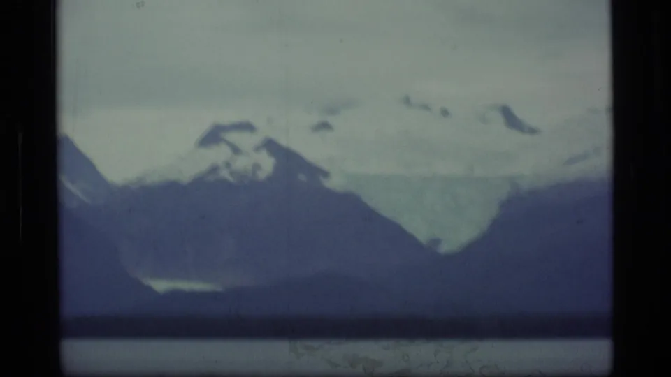 1977 Alaska 8mm frame with soft-focus snowy slopes, dark foreground mountains, and lake water below.