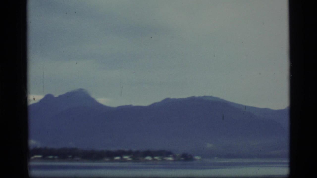 1977 Alaska 8mm frame showing misty mountains beyond calm water and small shoreline buildings.