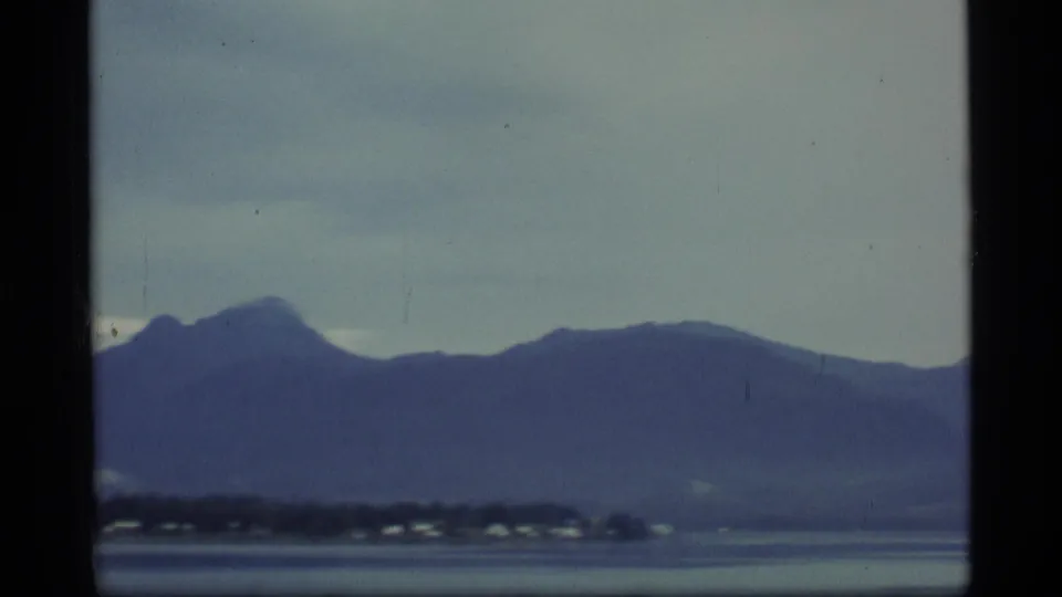 1977 Alaska 8mm frame showing misty mountains beyond calm water and small shoreline buildings.