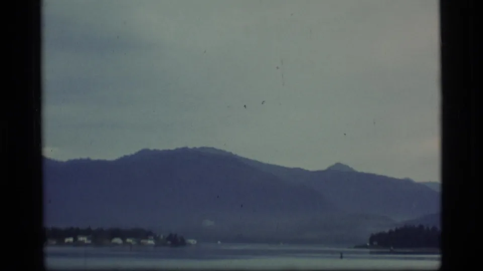 1977 Alaska home-movie view of a mountain range across water, with shoreline structures below the peaks.