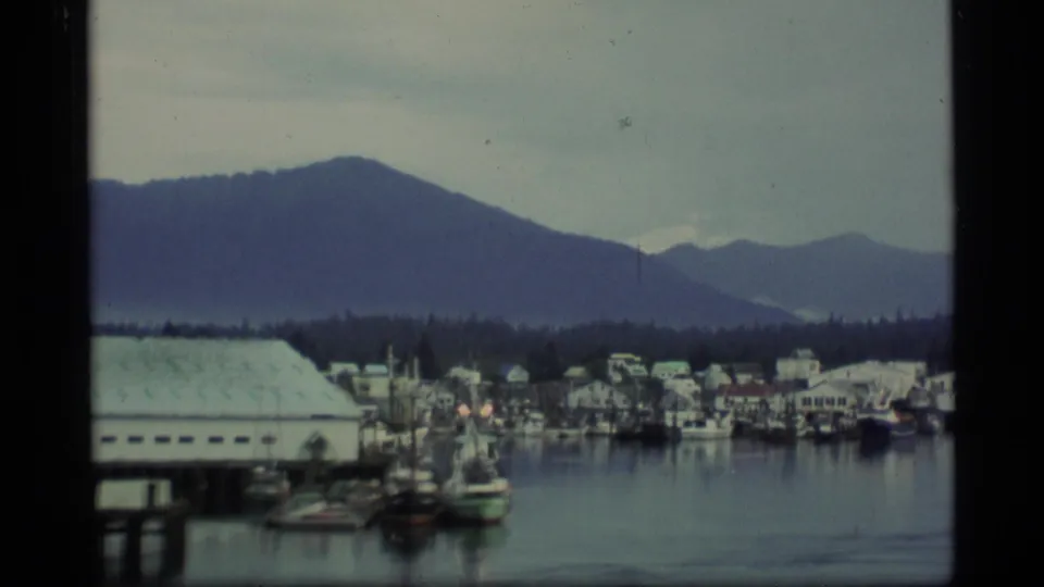 1977 Alaska 8mm frame showing a marina with boats, docks, waterfront buildings, and mountains behind.