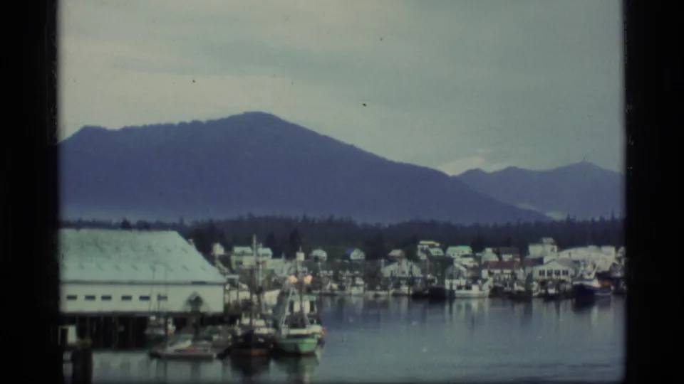 1977 Alaska home-movie frame of boats and harbor buildings set against misty mountains.
