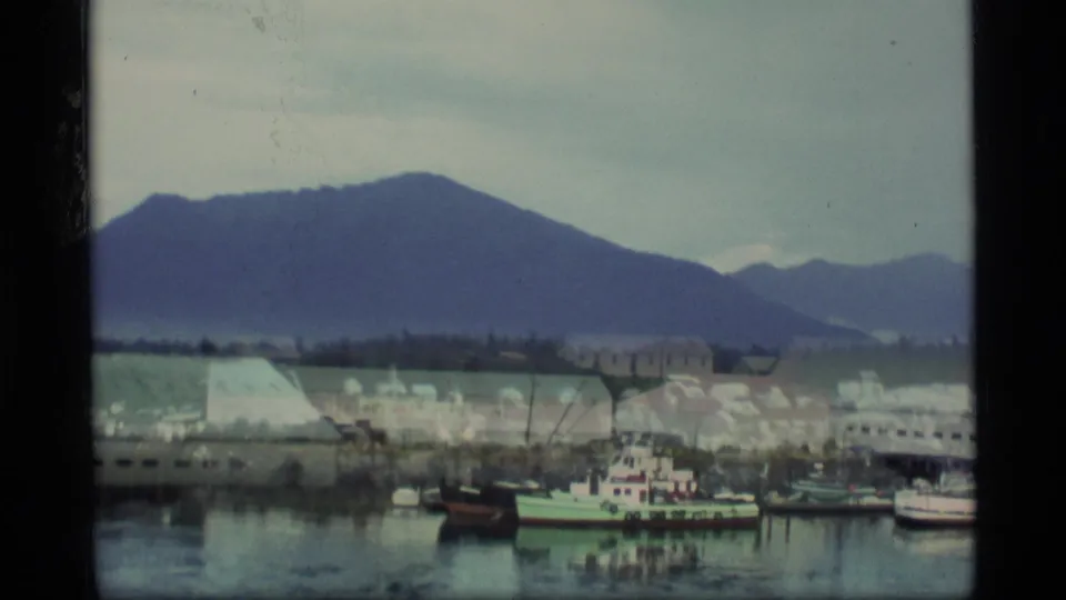 1977 Alaska home-movie view of a quiet fishing port with small boats, low waterfront buildings, and forested mountains under cloud cover.