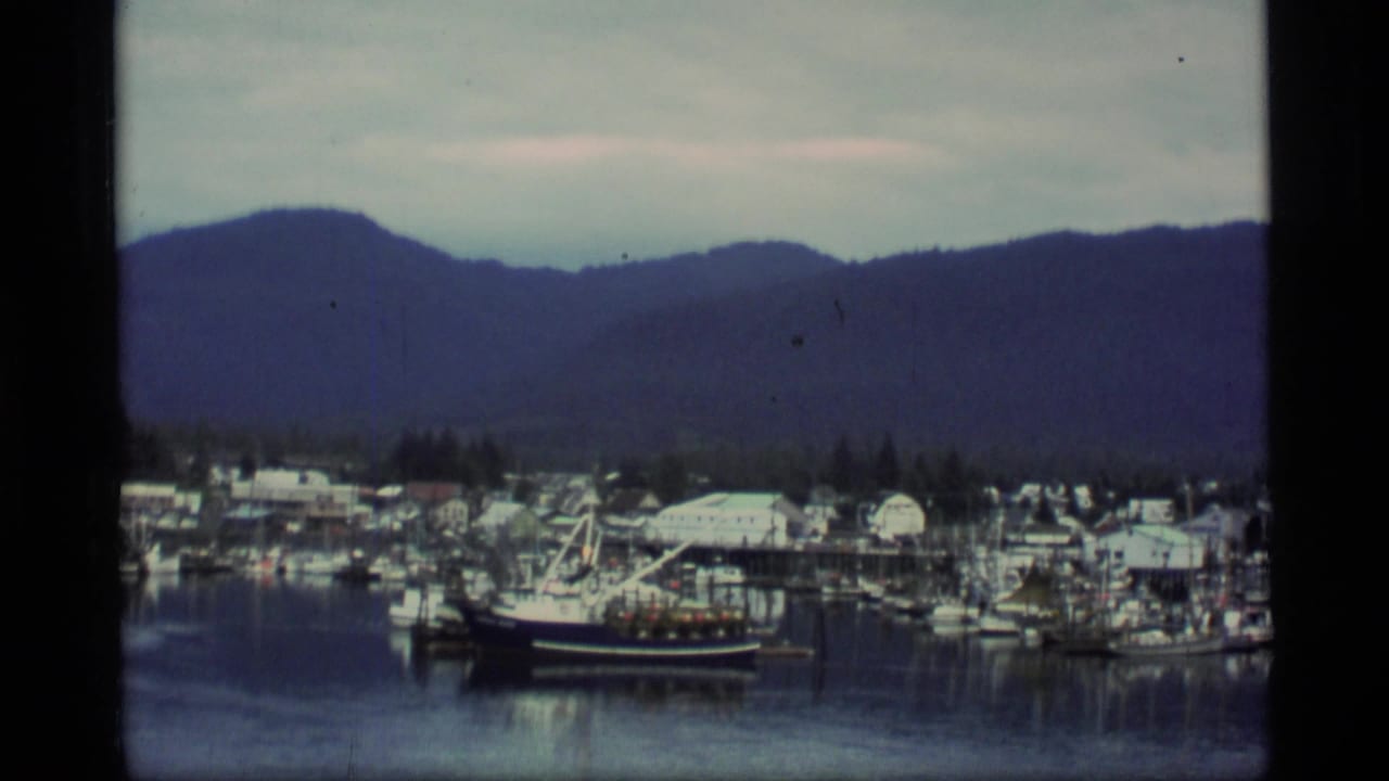 1977 Alaska harbor view with docked fishing boats, low shoreline buildings, calm water, and a dark mountain ridge beyond.