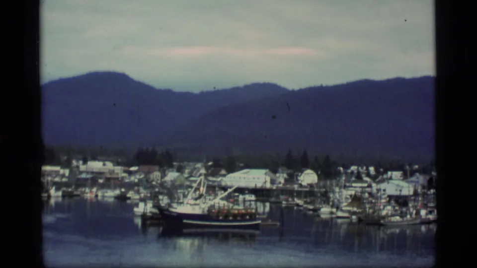 1977 Alaska harbor view with docked fishing boats, low shoreline buildings, calm water, and a dark mountain ridge beyond.