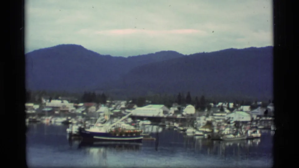 1977 Alaska marina scene with clustered boats, dock structures, waterfront buildings, trees, and mountains under a pale sky.