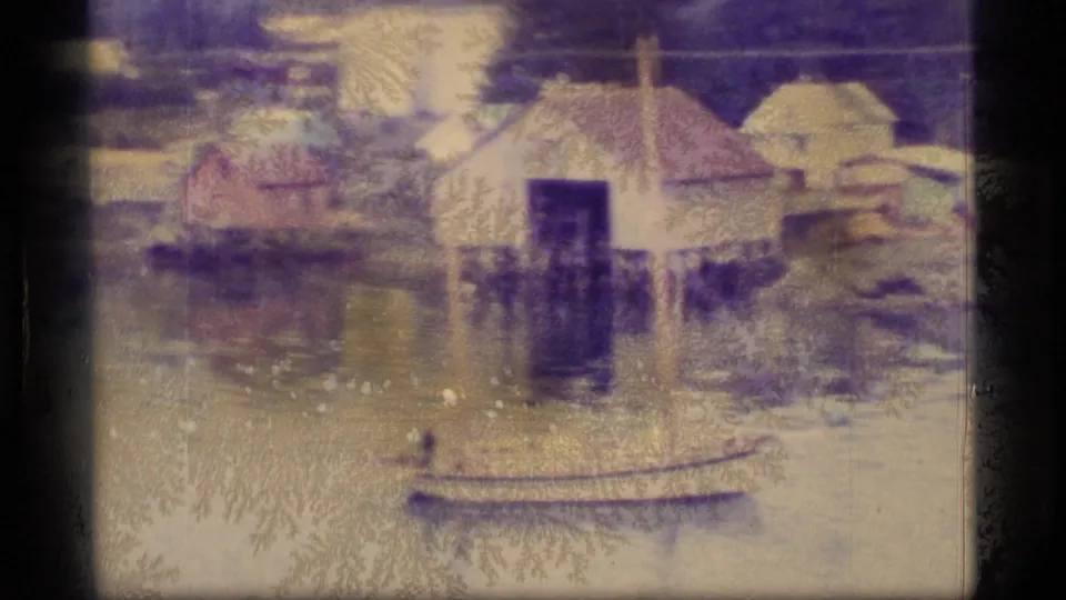 1977 Alaska waterfront detail showing buildings on pilings, reflective water, shoreline structures, and a small boat in the foreground.